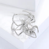 Spider brooch in polished silver-tone metal with large clear faceted teardrop center stone, side view on white block.