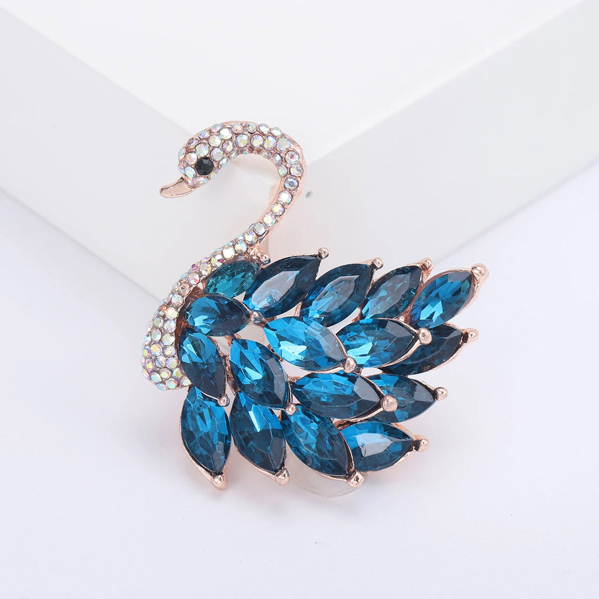 Swan brooch with rose gold-tone base, blue marquise-cut faceted stones, and pavé crystal head, front view and white studio setting.