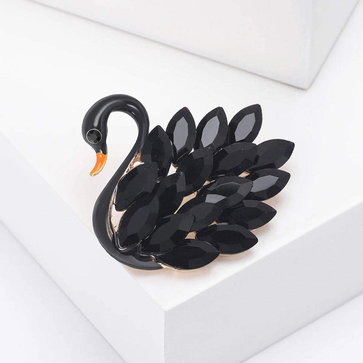 Swan brooch with black-tone body, orange beak, and marquise-cut black faceted stones, angled view on white block.