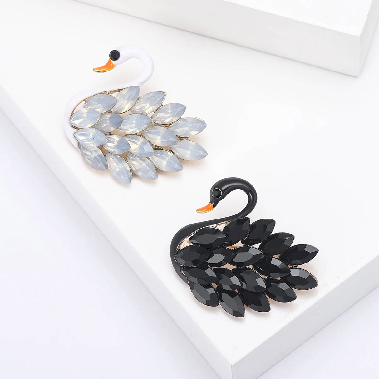 Pair of swan brooches with black and white enamel bodies and marquise-cut stones as feathers. Top-down view on white studio background.