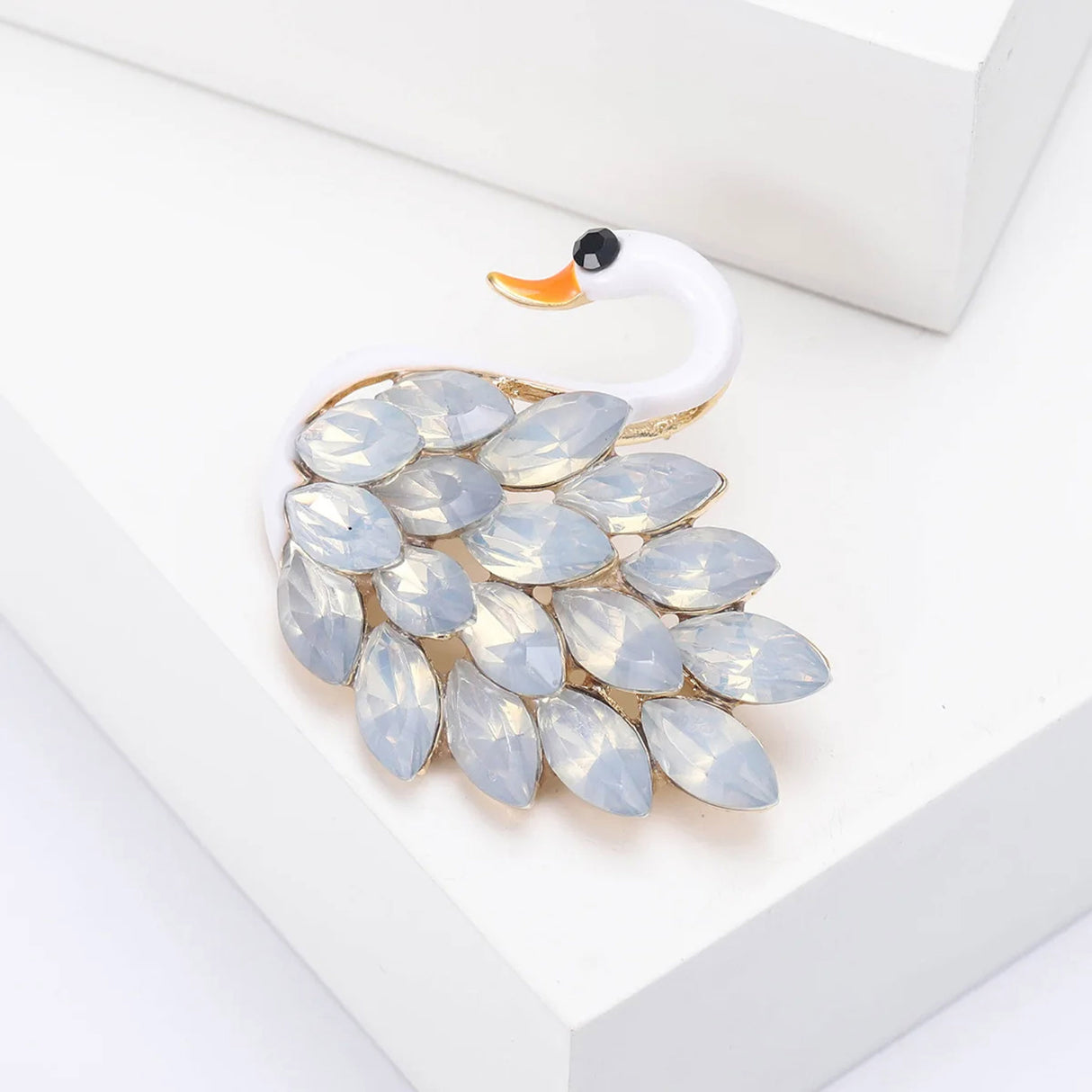 Swan brooch with white-tone body, orange beak, and white iridescent marquise-cut stones, angled view on white block.