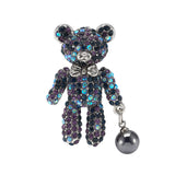 Teddy bear brooch with purple and blue iridescent stones, silver-tone bow, and dark metallic hanging faux pearl, cutout on white.