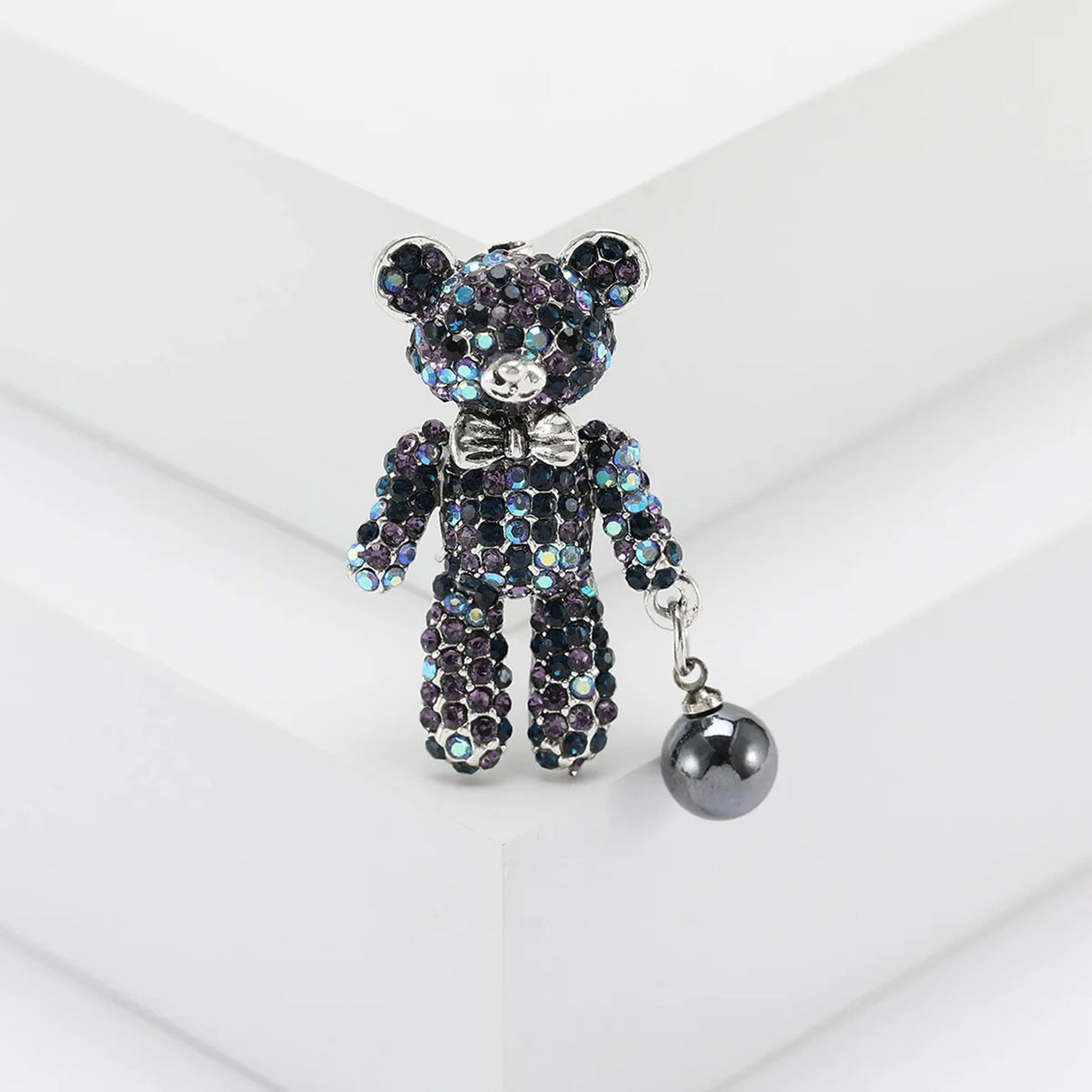 Teddy bear brooch with iridescent multicolor blue stones, silver-tone bow, and black hanging bead, upright on white block.