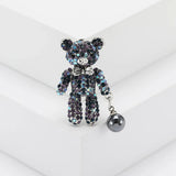 Teddy bear brooch with iridescent multicolor blue stones, silver-tone bow, and black hanging bead, upright on white block.