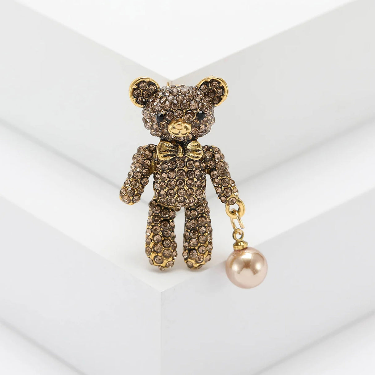 Teddy bear brooch with brown-tone stones, gold-tone bow, and light bronze hanging orb, upright pose on white platform.