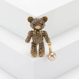 Teddy bear brooch with brown-tone stones, gold-tone bow, and light bronze hanging orb, upright pose on white platform.