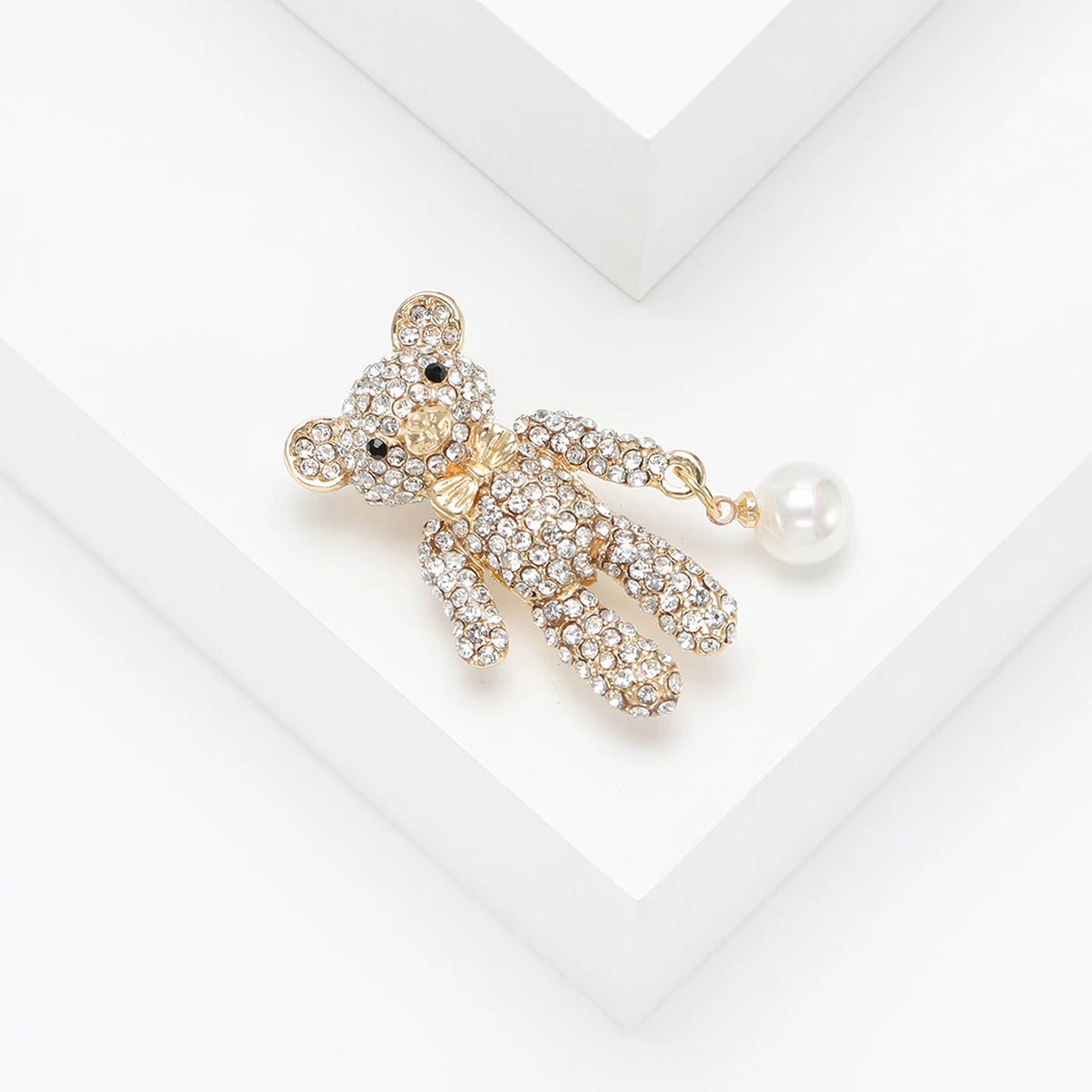 Teddy bear brooch with clear stones, gold-tone details, and white dangling bead, angled top-down view on white.