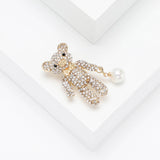 Teddy bear brooch with clear stones, gold-tone details, and white dangling bead, angled top-down view on white.