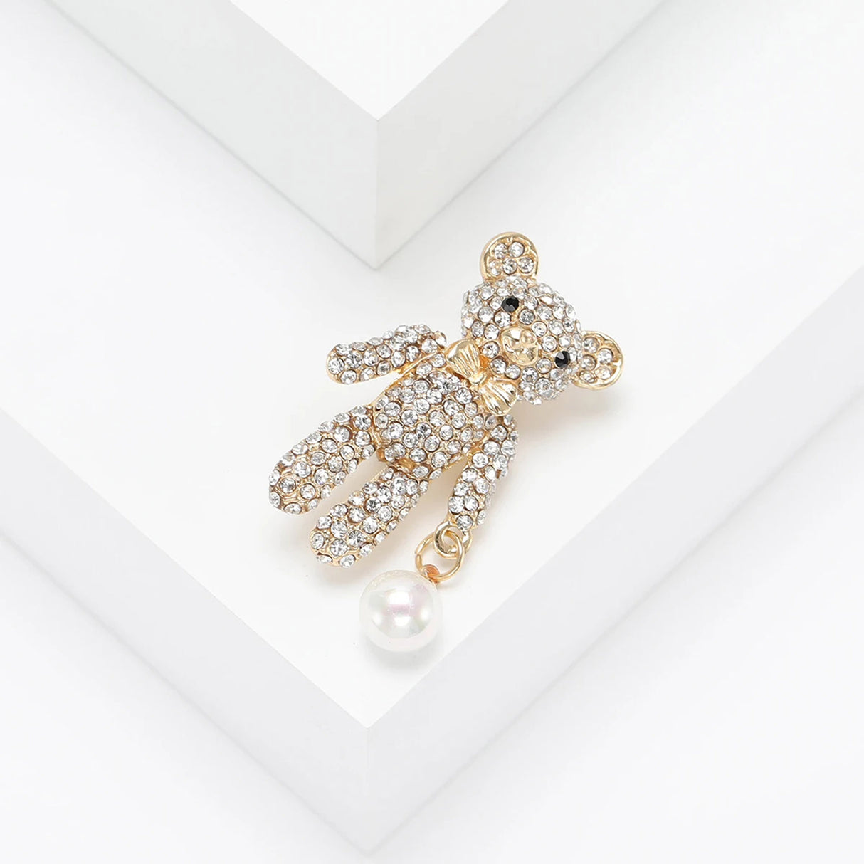 Teddy bear brooch with clear faceted stones, gold-tone bow, and white round bead charm, top-right view on white surface.