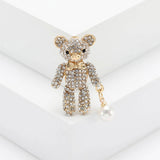 Teddy bear brooch in gold-tone metal with clear faceted stones and white imitation pearl charm, upright on white block.