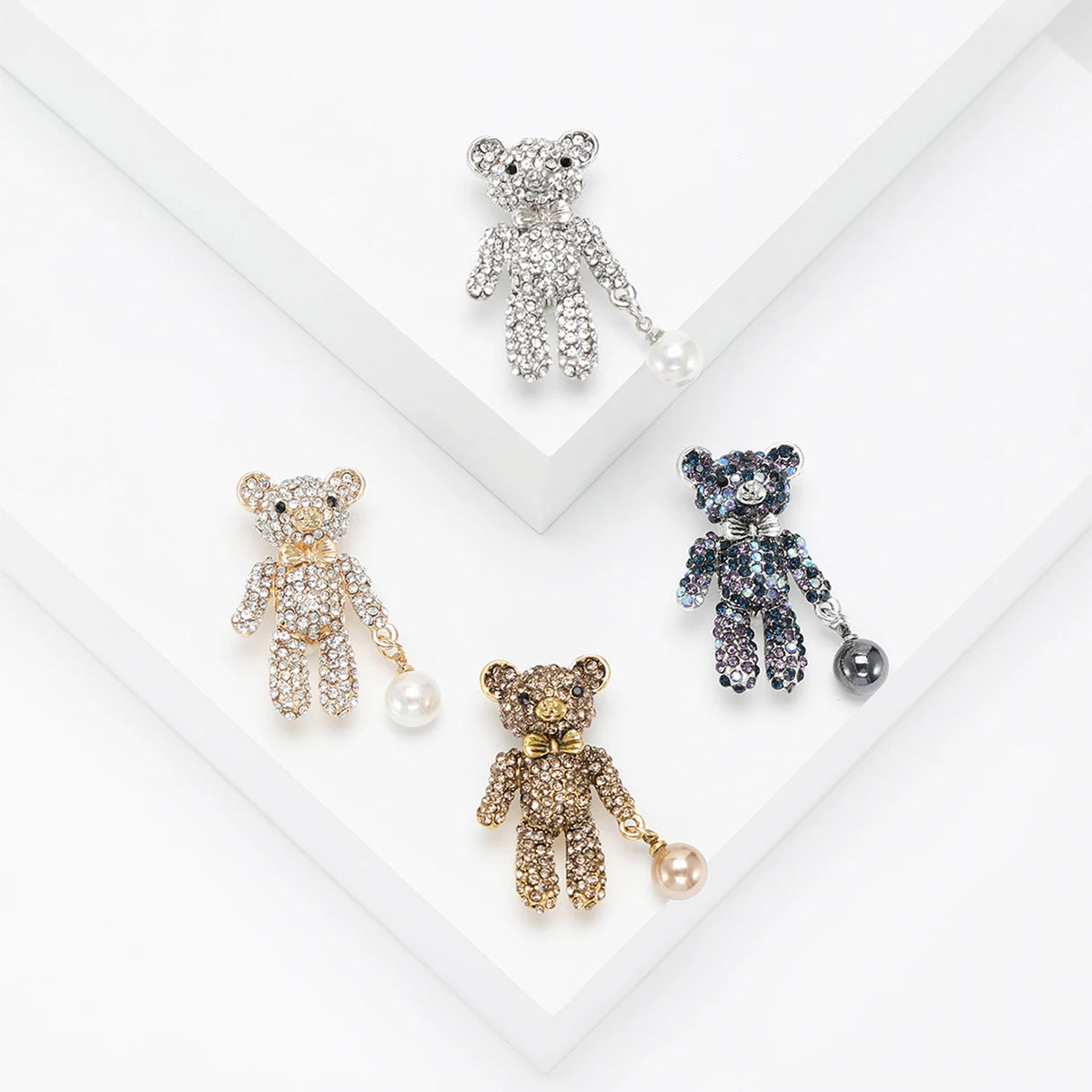 Set of four teddy bear brooches in silver, gold, and blue tone finishes with matching dangling beads, flat lay on white steps.
