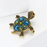 Turtle brooch with gold-tone textured surface and blue-green decorative stones, angled view on white surface.