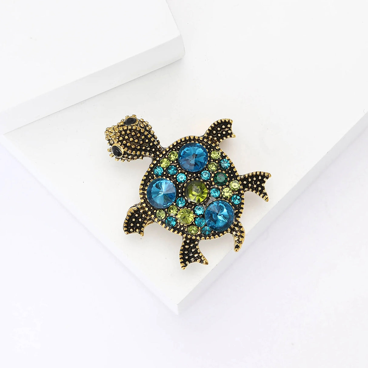Turtle brooch with gold-tone metal finish and assorted blue and green stones, flat lay on white background.