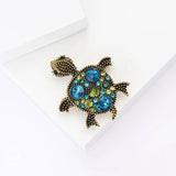 Turtle brooch with gold-tone metal finish and assorted blue and green stones, flat lay on white background.