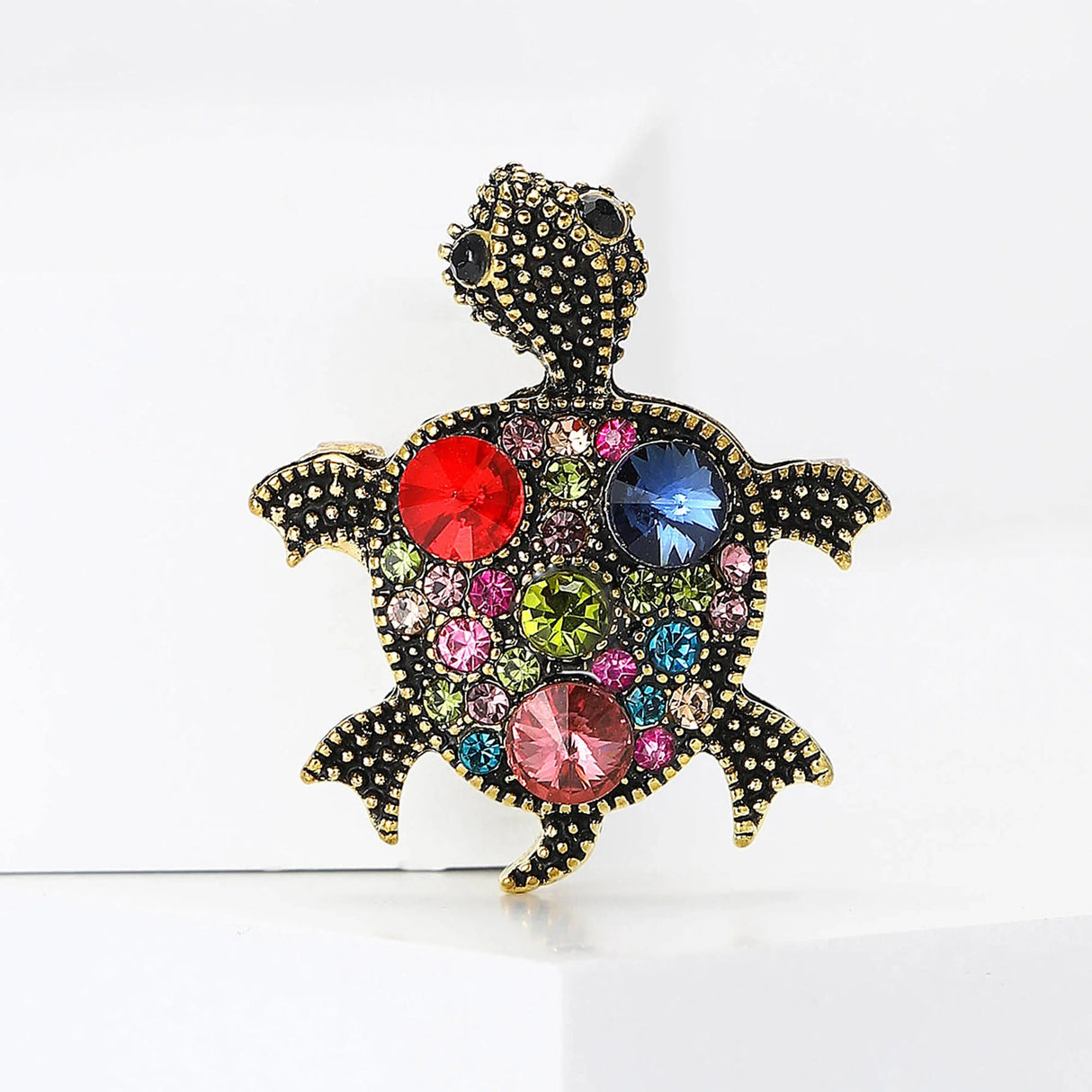 Vintage-inspired turtle brooch with gold-tone dotted metal texture and multicolor faceted stones in red, pink, blue, and green, upright pose on white studio backdrop.