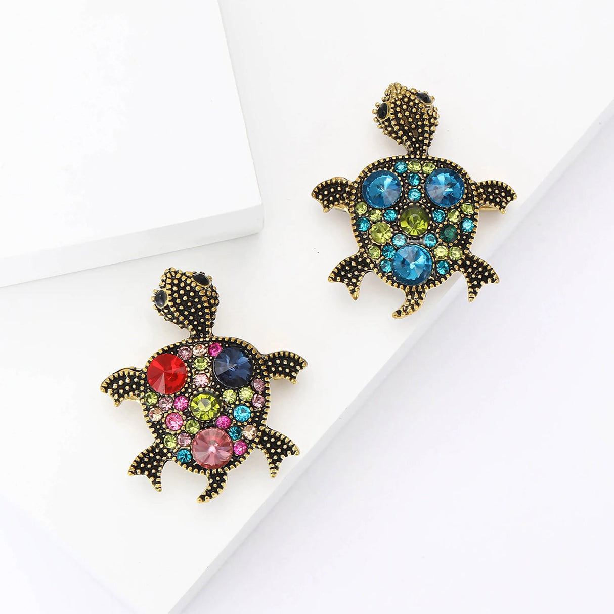 Pair of turtle brooches with gold-tone dotted metal finish, one with multicolor stones and one with blue-green stones, flat lay on white.