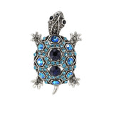 Tortoise brooch with silver-tone metal finish, dark blue center stones, and light blue faceted accents, cutout on white.