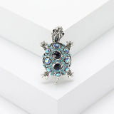 Tortoise brooch with silver-tone metal frame, dark blue faceted center stones, and light blue accents, standing on white stepped platform.