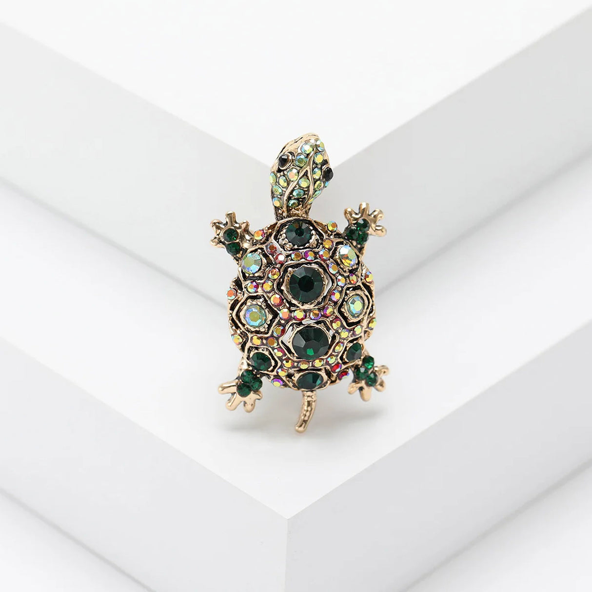 Tortoise brooch with gold-tone metal base, dark green faceted stones, and iridescent accents, standing on white stepped platform.