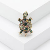 Tortoise brooch with gold-tone metal base, dark green faceted stones, and iridescent accents, standing on white stepped platform.