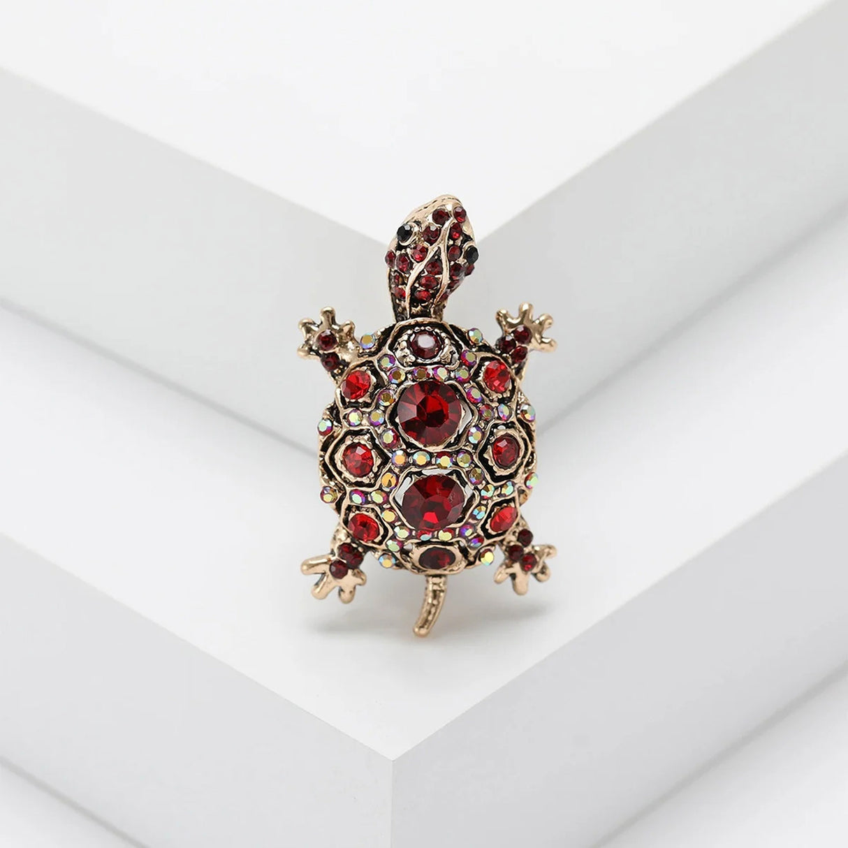 Tortoise brooch with gold-tone metal base, red faceted center stones, and smaller iridescent red accents, standing on white stepped studio platform.
