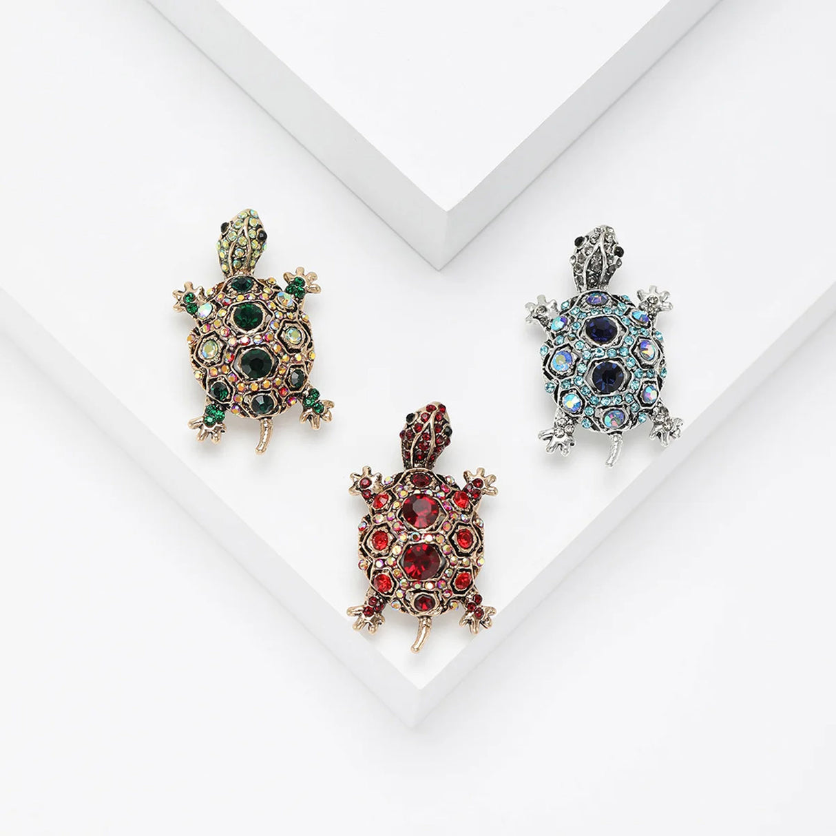 Set of three tortoise brooches in gold and silver-tone finishes with red, green, and blue faceted stones, flat lay on white stepped surface in studio.