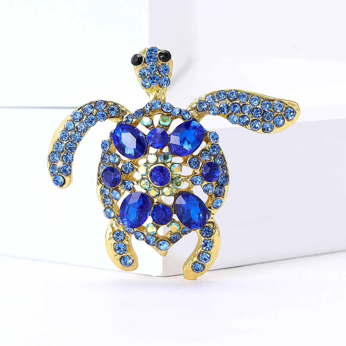 Sea turtle brooch with blue stones and gold-tone metal frame, displayed upright against white block in studio.