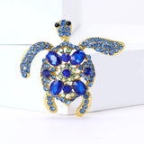 Sea turtle brooch with blue stones and gold-tone metal frame, displayed upright against white block in studio.