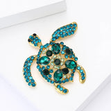 Turtle brooch with teal stones, darker green accents, and gold-tone metal finish. Angled top-down view on white studio surface.