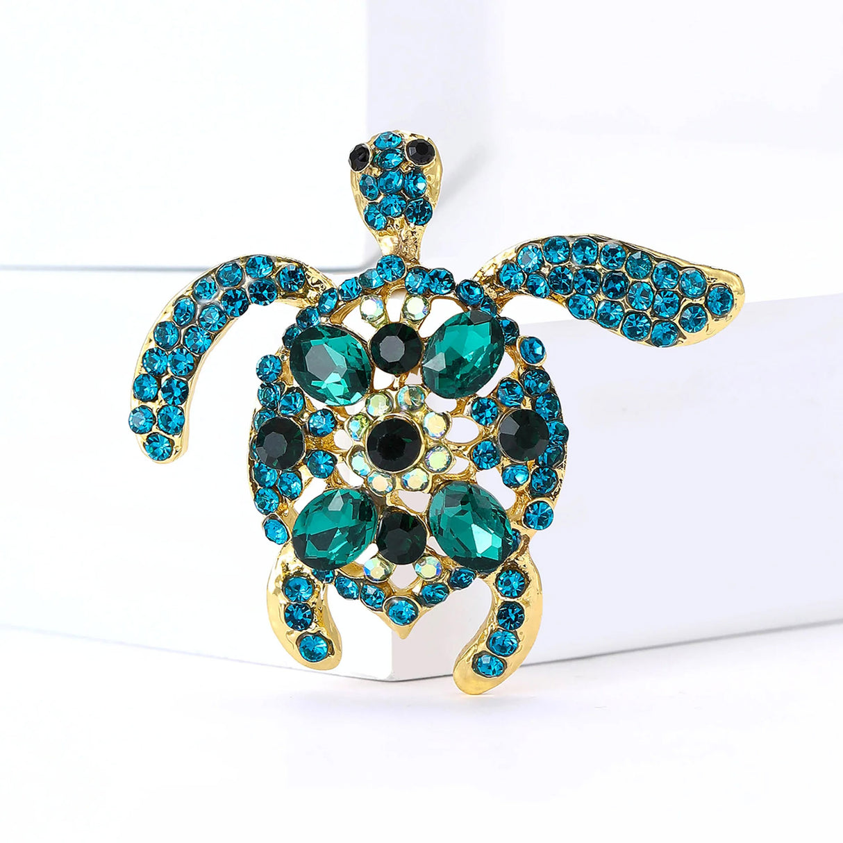 Turtle brooch with teal and green faceted stones, set in gold-tone metal body, standing on white block.