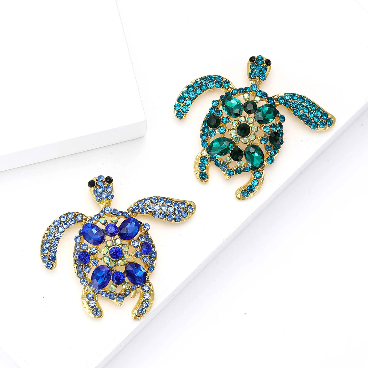 Pair of sea turtle brooches with gold-tone metal frame, one in blue stones and the other in teal stones, top-down view on white studio backdrop.