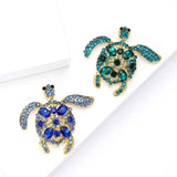 Pair of sea turtle brooches with gold-tone metal frame, one in blue stones and the other in teal stones, top-down view on white studio backdrop.