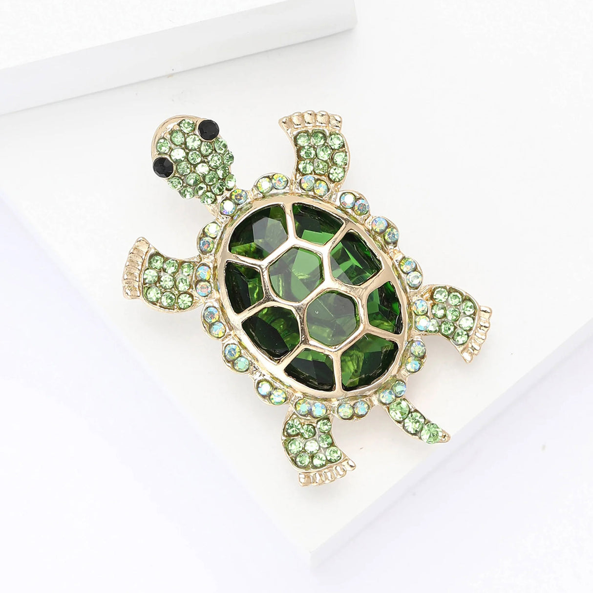 Turtle brooch with deep green faceted shell stone, light green round accent stones, and gold-tone trim, angled view on white block.
