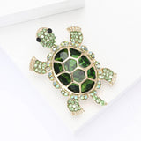 Turtle brooch with deep green faceted shell stone, light green round accent stones, and gold-tone trim, angled view on white block.