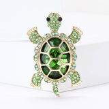 Turtle brooch featuring deep green center stone, pale green round accents, and gold-tone metal, standing on white surface.