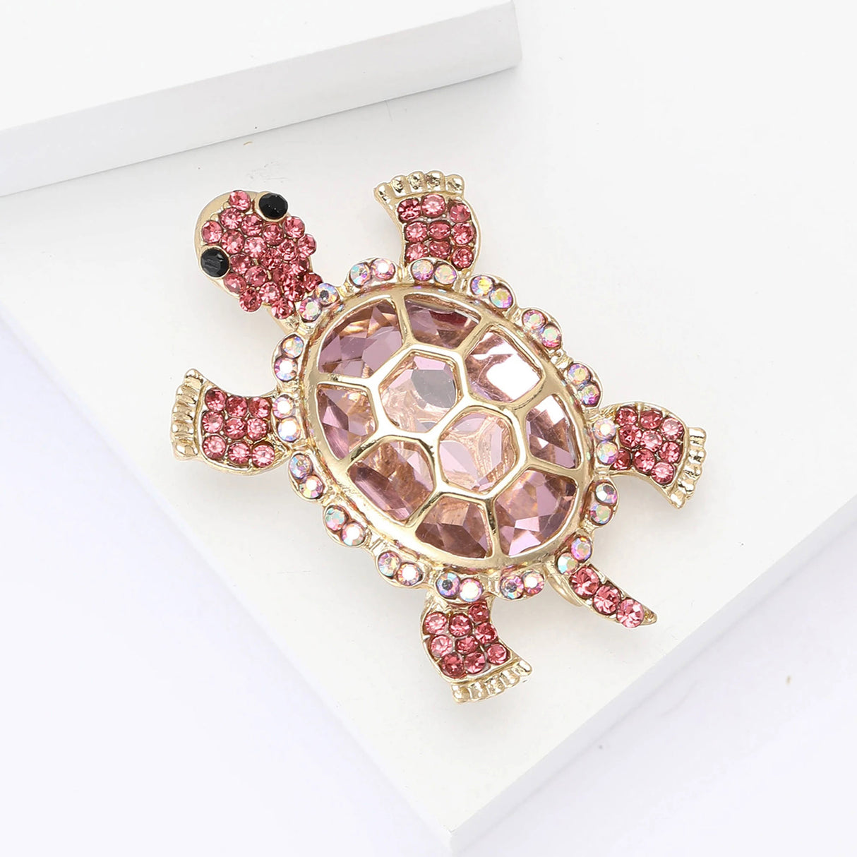Turtle brooch with pink faceted center stone, vivid pink round accents, and gold-tone setting, angled view on white block.