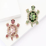 Pair of tortoise brooches with pink and green faceted center stones and small round accents, set in gold-tone metal. Flat lay on white blocks in studio.