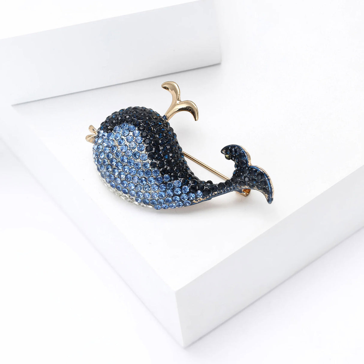 Whale brooch featuring blue faceted stones with gold-tone accents, angled side view on white block.