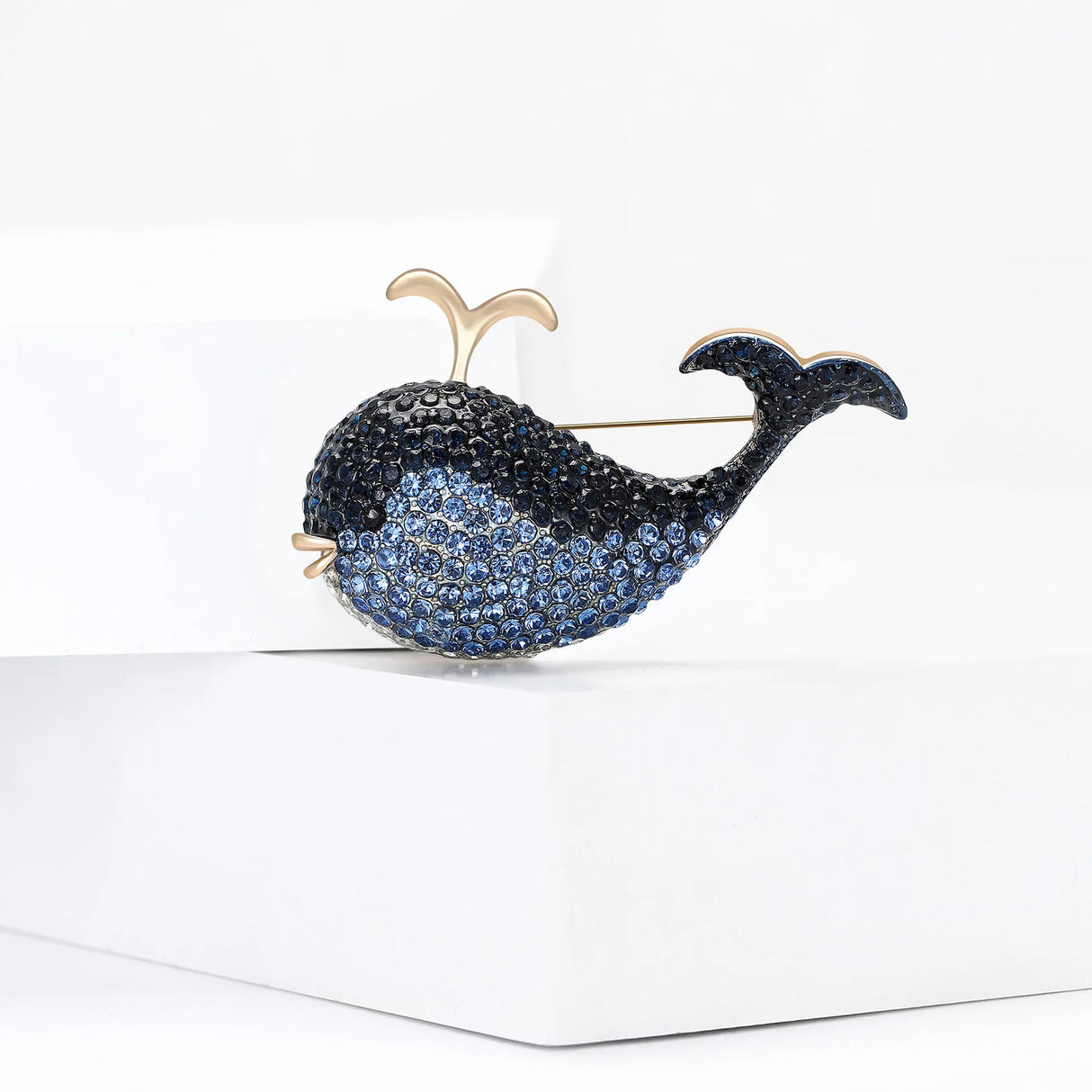 Whale brooch covered in blue faceted stones with gold-tone spout detail, front view on white platform.